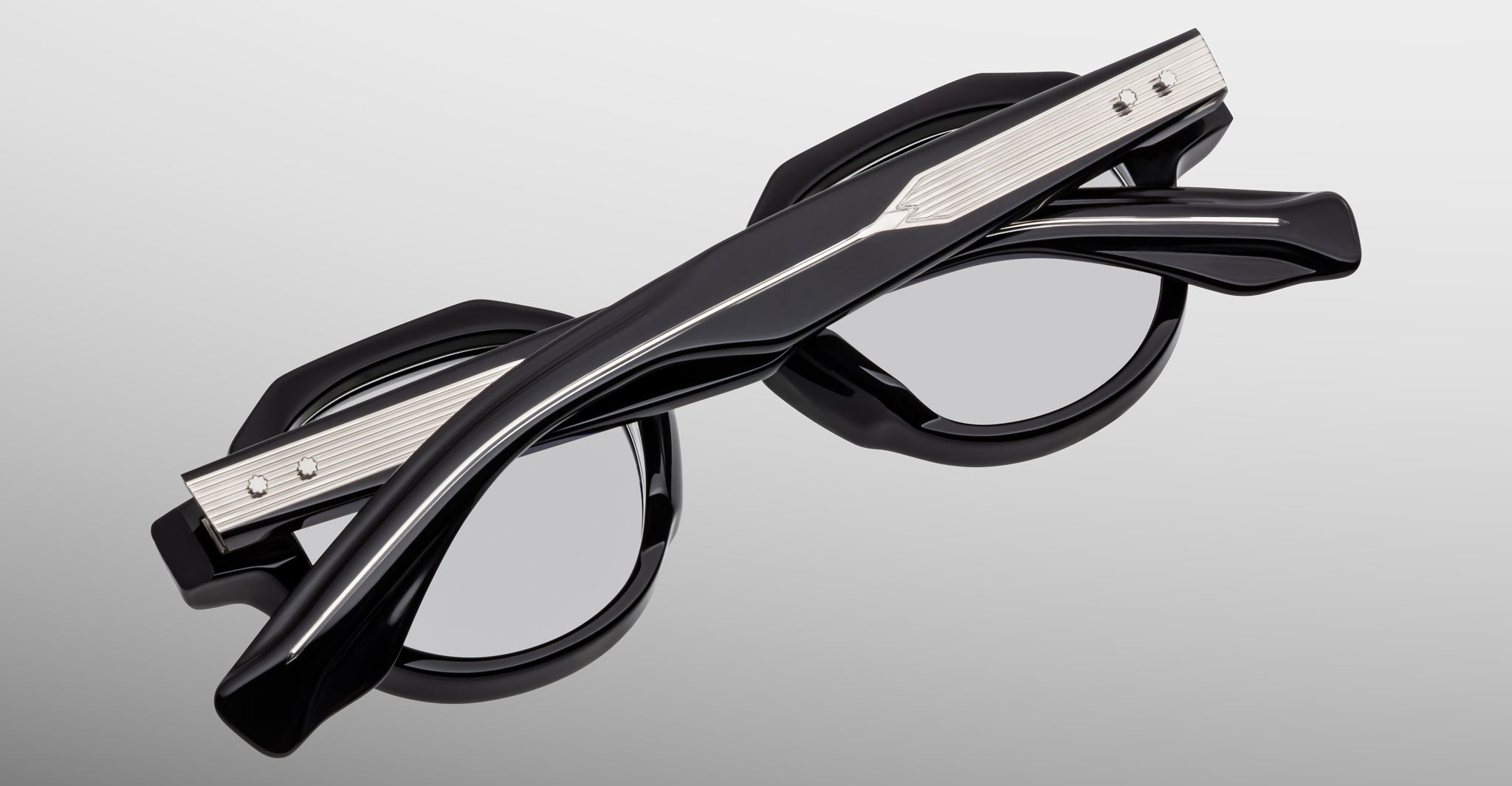 The Jacques Marie Mage Pontian is a pair of black round eyeglasses with thick, angular arms featuring metallic silver details, shown folded with crossed arms on a light gray background.