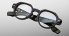 A pair of Jacques Marie Mage Pontian black, thick-rimmed eyeglasses with round lenses and silver hinge accents rests on a light gray background.