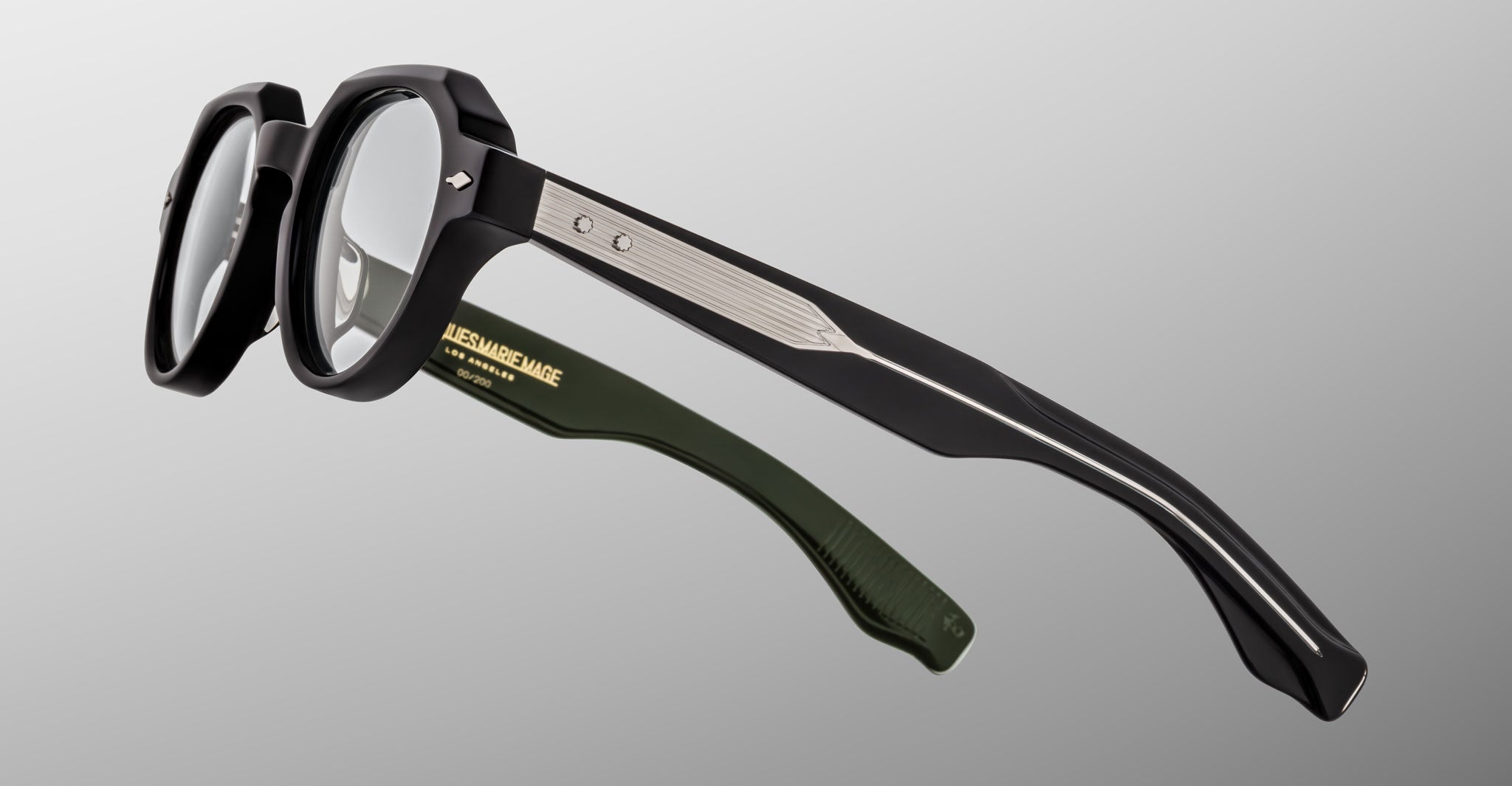 The Jacques Marie Mage Pontian eyeglasses feature black frames, clear lenses, silver temple accents, and green inner temple arms, shown on a light gray background.