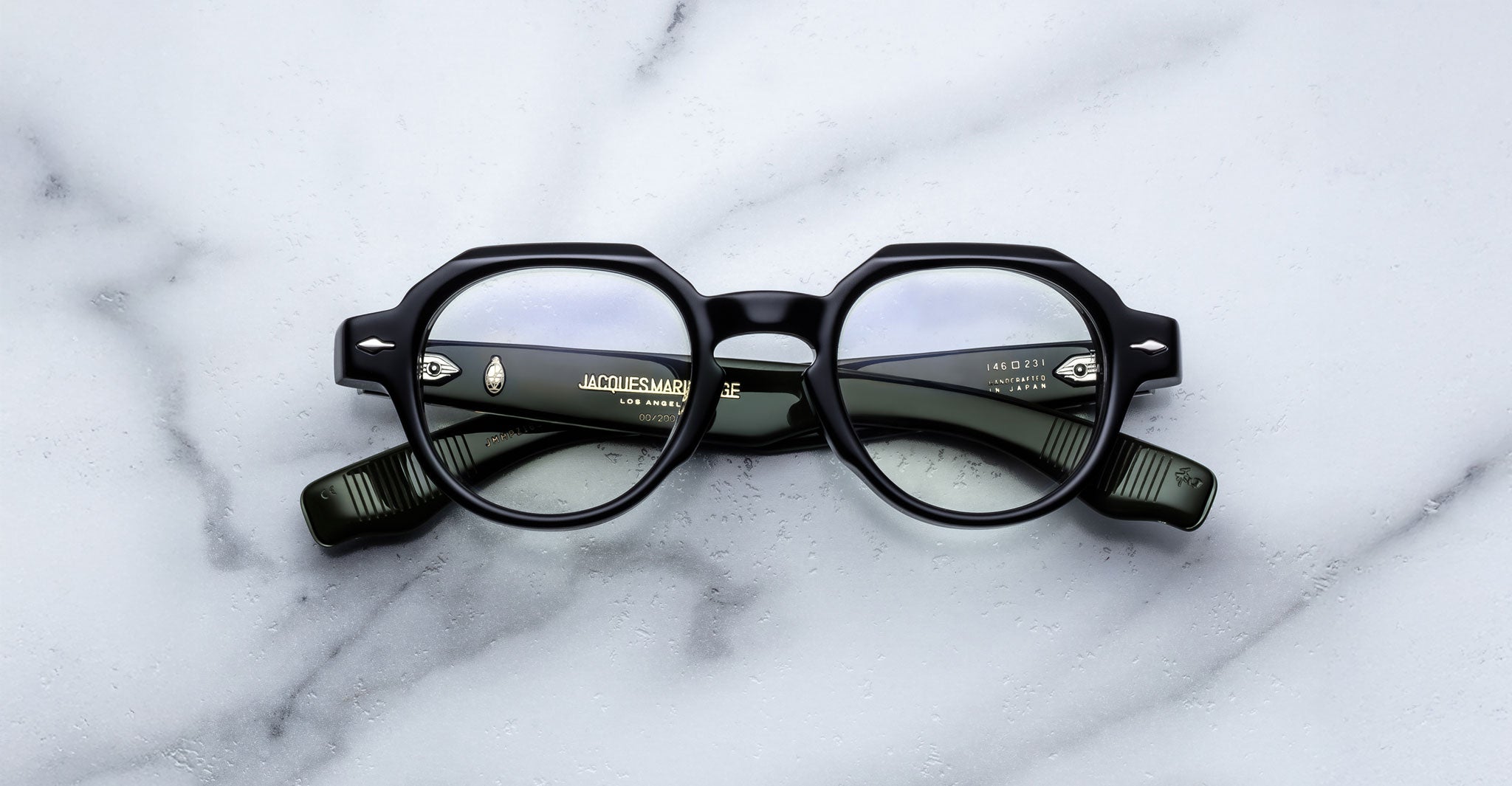 The Jacques Marie Mage Pontian is a pair of round black eyeglasses with thick frames and green arms, resting on a white marble surface. Brand information appears in white text inside the left arm.