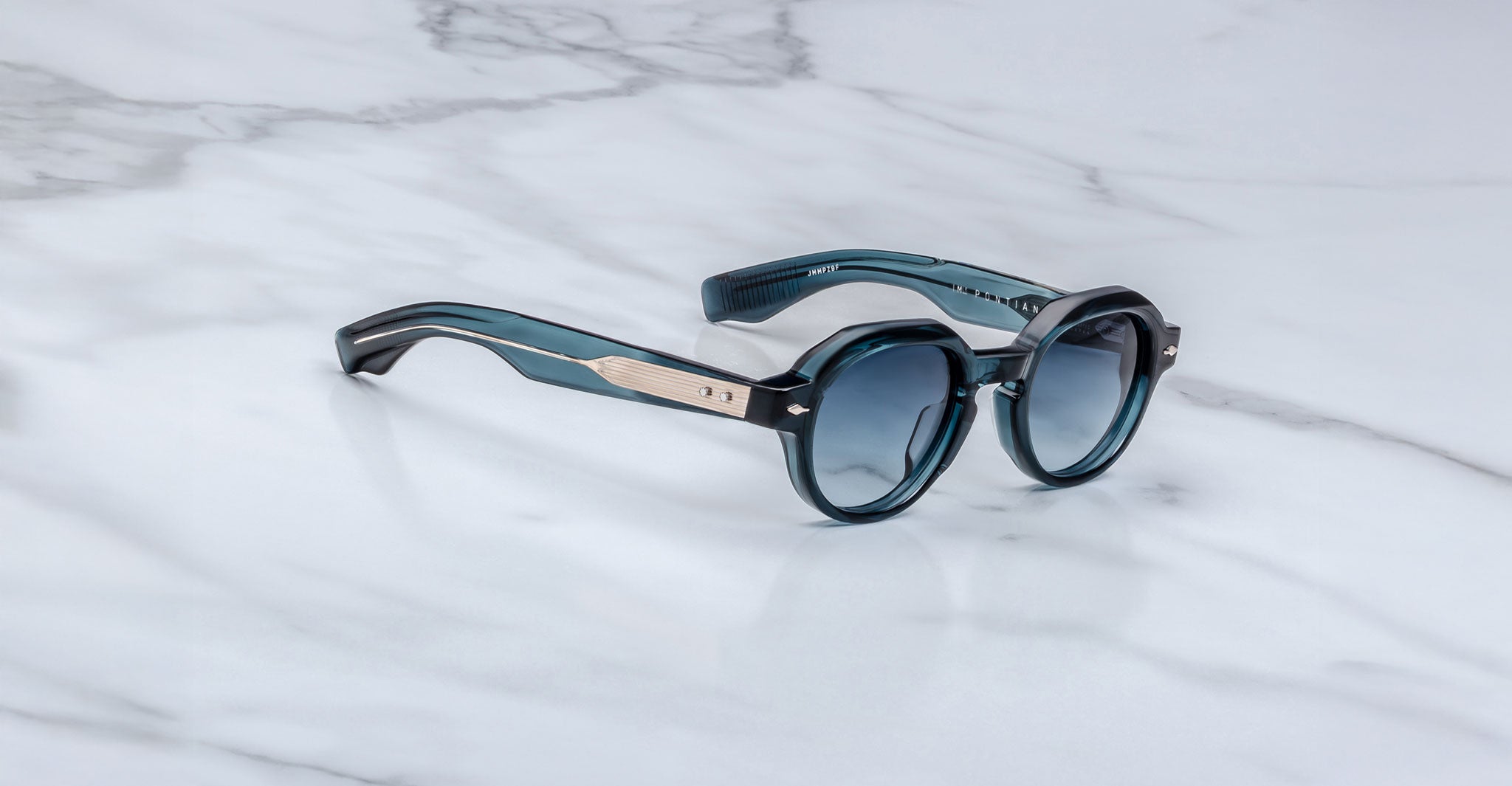 The Jacques Marie Mage Pontian sunglasses feature round, blue-tinted lenses, thick dark blue frames, and metallic temple accents, displayed on a white marble surface.
