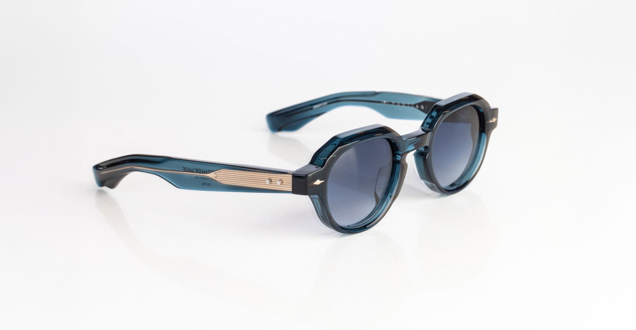 The Jacques Marie Mage Pontian sunglasses feature blue acetate round frames with dark-tinted lenses, beige metallic strip accents on the arms, and arrow-shaped details near the hinges, shown resting on a white surface.