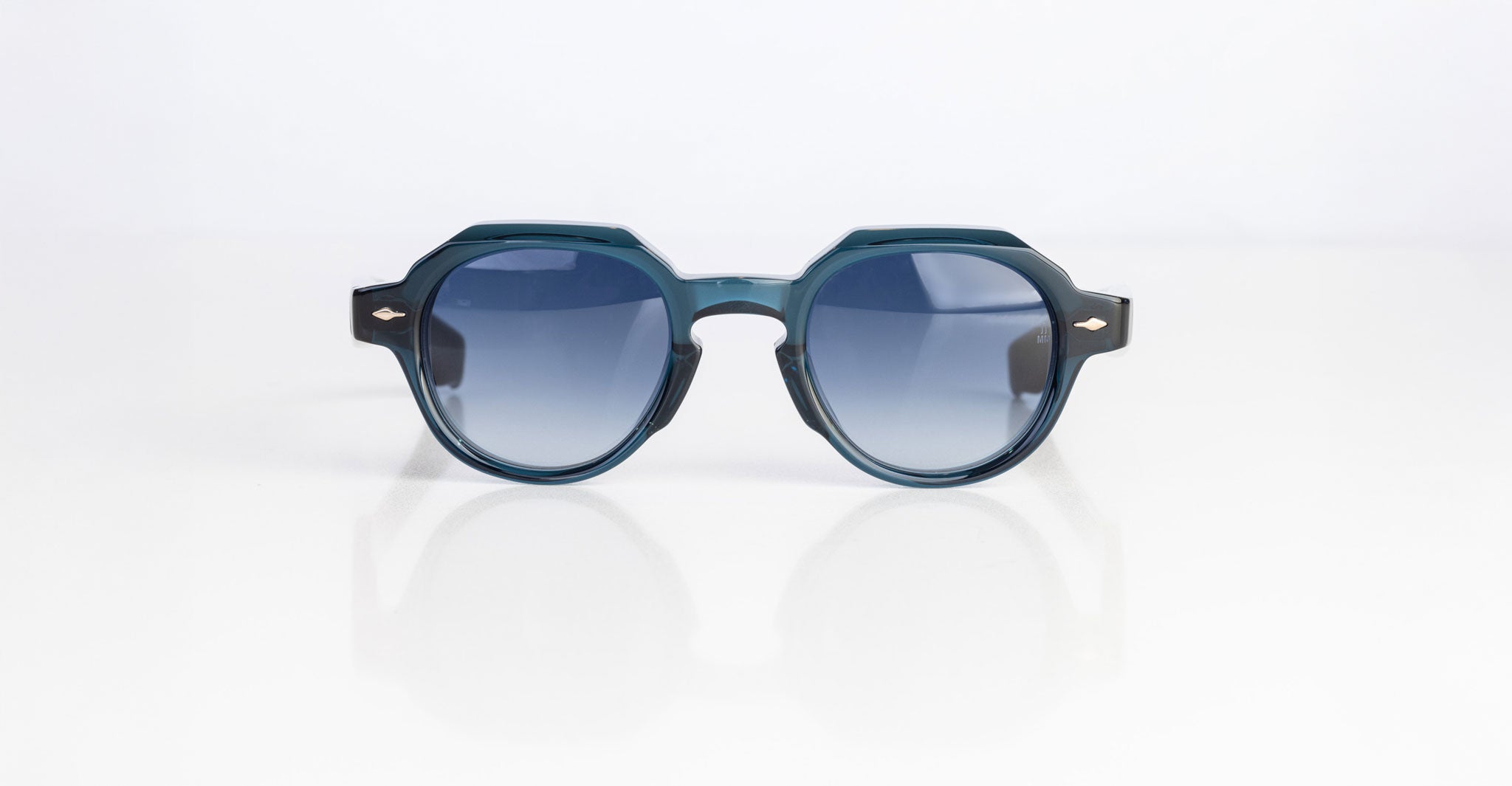 The Jacques Marie Mage Pontian sunglasses, featuring a round silhouette in dark teal with blue-tinted lenses, rest on a white surface against a plain white background.