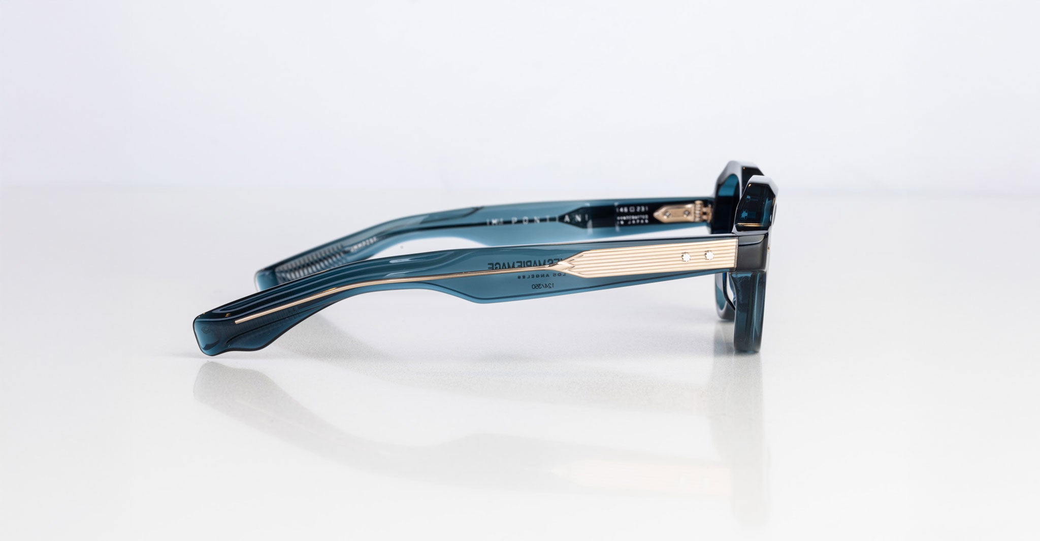 The Jacques Marie Mage Pontian eyeglasses, featuring a teal frame and gold-detailed temples, are displayed on a white reflective surface in a side-view photograph.