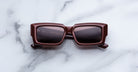 Jacques Marie Mage Quant in Bordeaux Empire, rectangular burgundy acetate sunglasses with bold sculpted rims, wide temples, and softly tinted lenses, shown front-facing on a light marble surface.