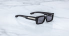 Jacques Marie Mage Quant in Shadow, rectangular black acetate sunglasses shown from a side angle, featuring bold sculpted rims, wide temples with signature metal inlay, and dark tinted lenses on a light marble surface.