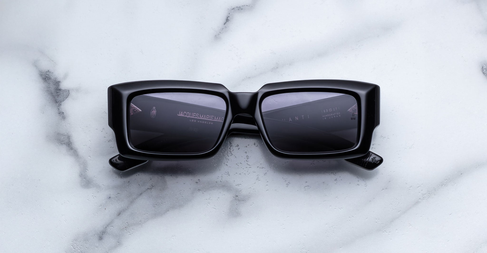 Jacques Marie Mage Quant in Shadow, rectangular black acetate sunglasses with bold sculpted rims, wide temples, and dark tinted lenses, shown front-facing on a light marble surface.
