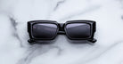 Jacques Marie Mage Quant in Shadow, rectangular black acetate sunglasses with bold sculpted rims, wide temples, and dark tinted lenses, shown front-facing on a light marble surface.