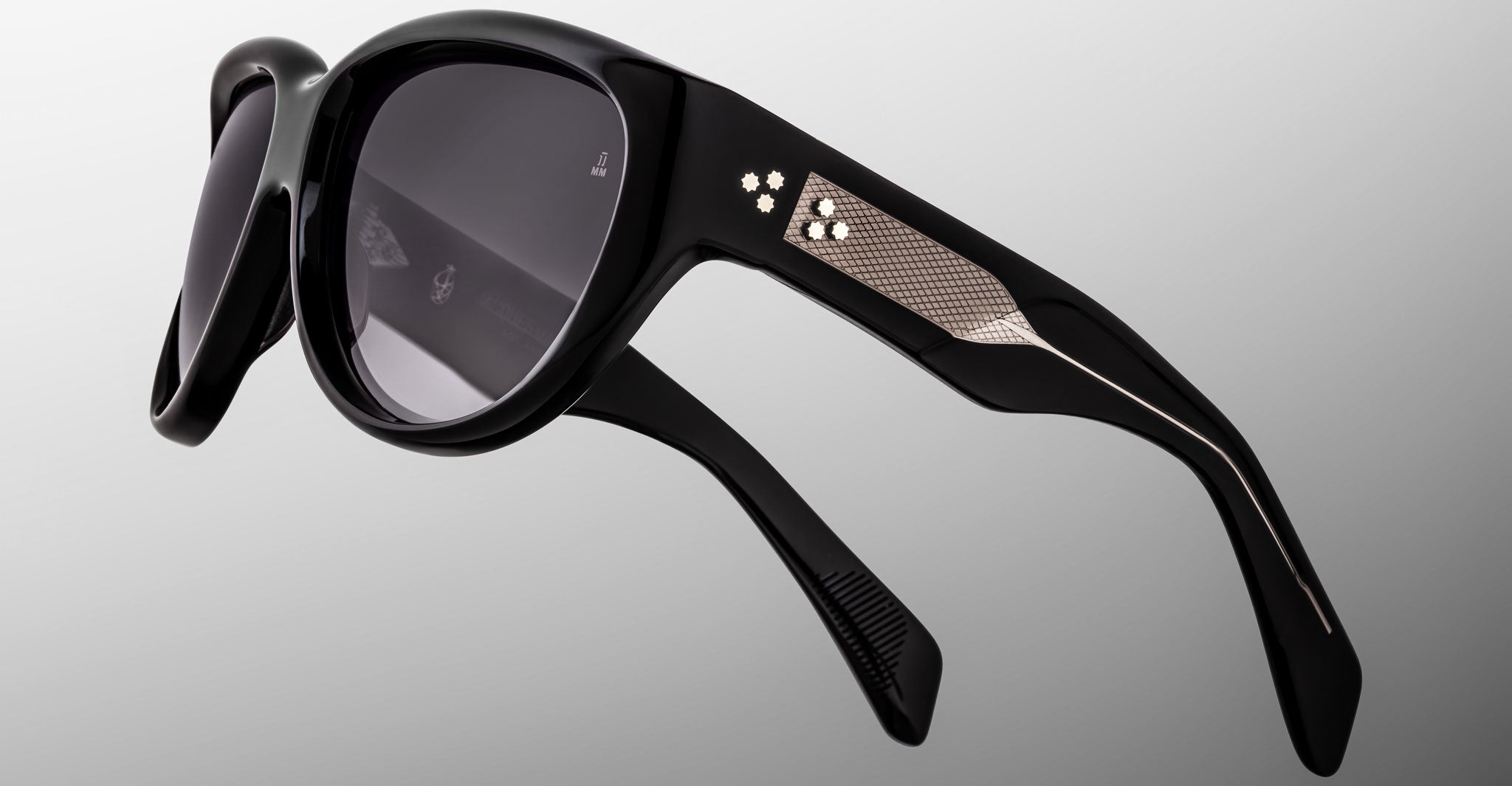 Close-up of Jacques Marie Mage Ravello black sunglasses with round dark lenses and textured metallic temple detail, featuring subtle decorative elements and a reflective finish, set against a light gray background.