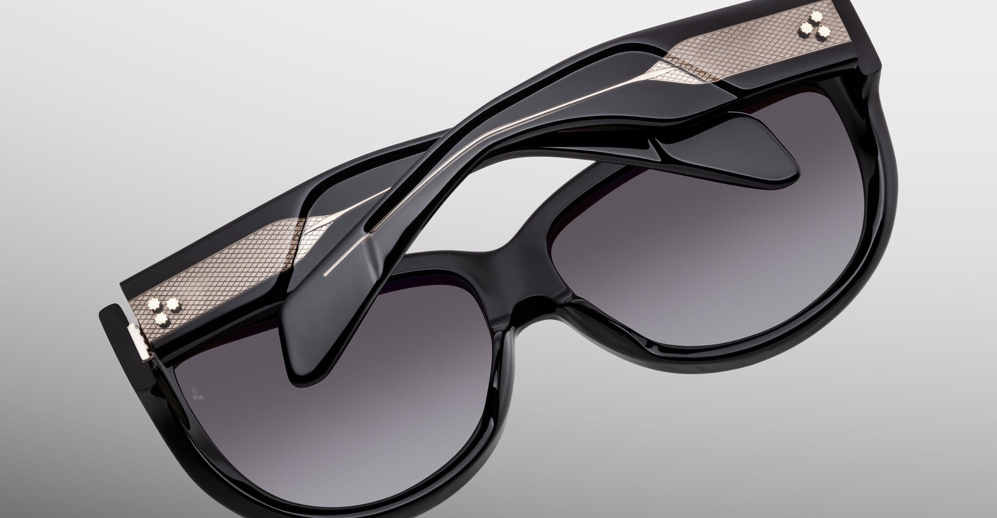 The Jacques Marie Mage Ravello sunglasses feature black frames, dark gradient lenses, metallic gold accents, and layered sculptural temples, shown against a plain gray background.