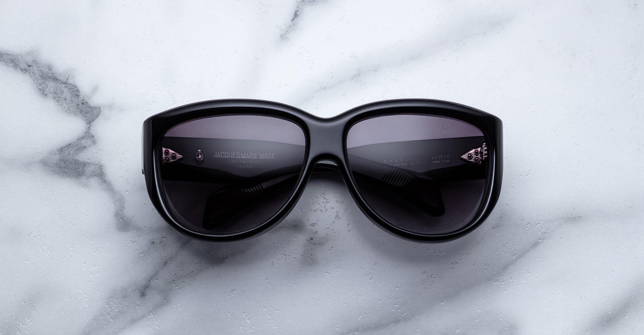 A Jacques Marie Mage Ravello pair of black sunglasses with dark lenses is placed on a white marble surface with grey veining, the lenses facing upward.