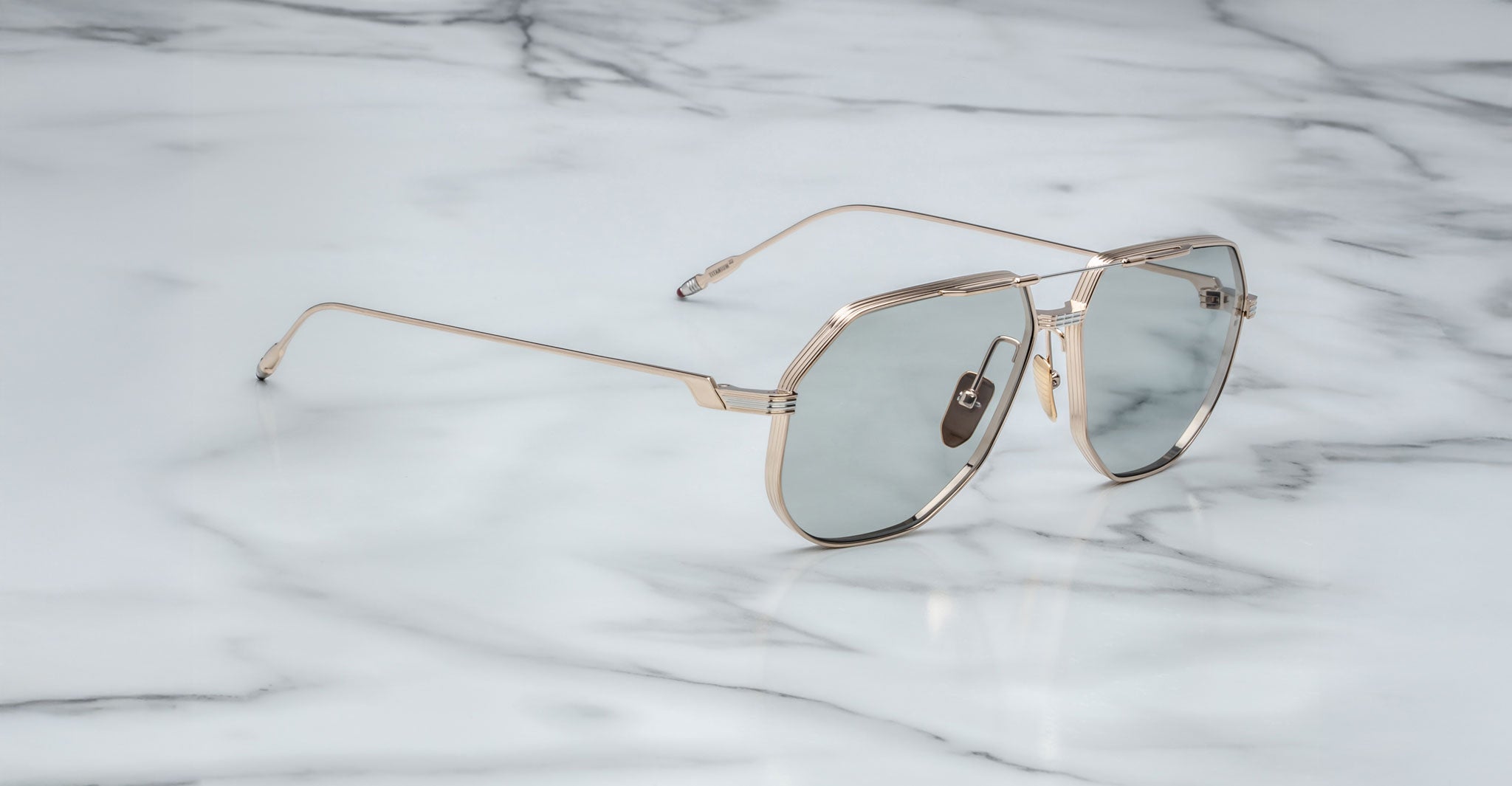 The Jacques Marie Mage Reynold sunglasses, featuring stylish oversized aviator frames in thin gold metal with light-tinted lenses, rest on a white marble surface with subtle gray veining, adding vintage Americana charm.