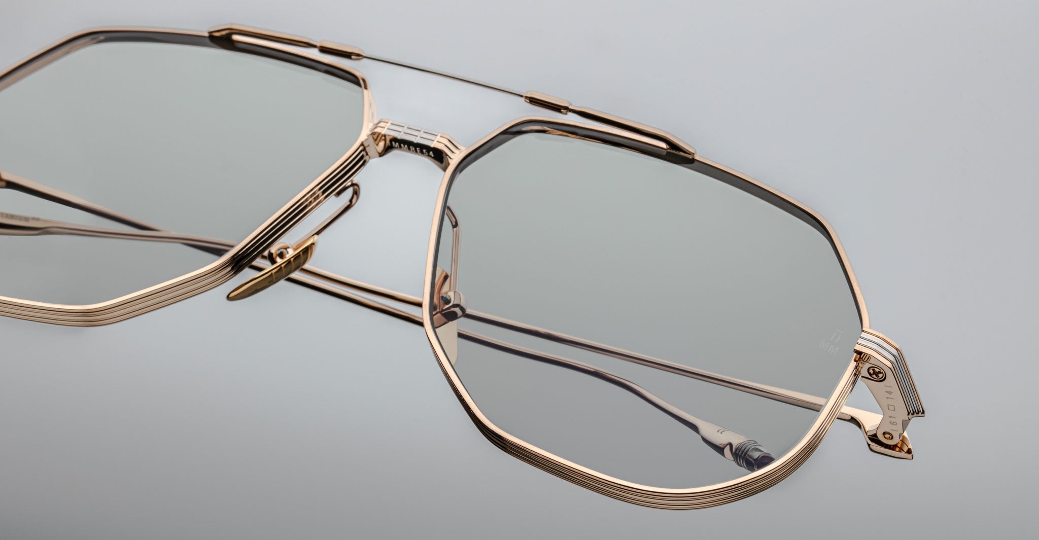 Close-up of Jacques Marie Mage Reynold eyeglasses featuring a gold-rimmed, geometric titanium frame with fine detailed lines, slightly tinted lenses, and clear nose pads, set against a light gray background.