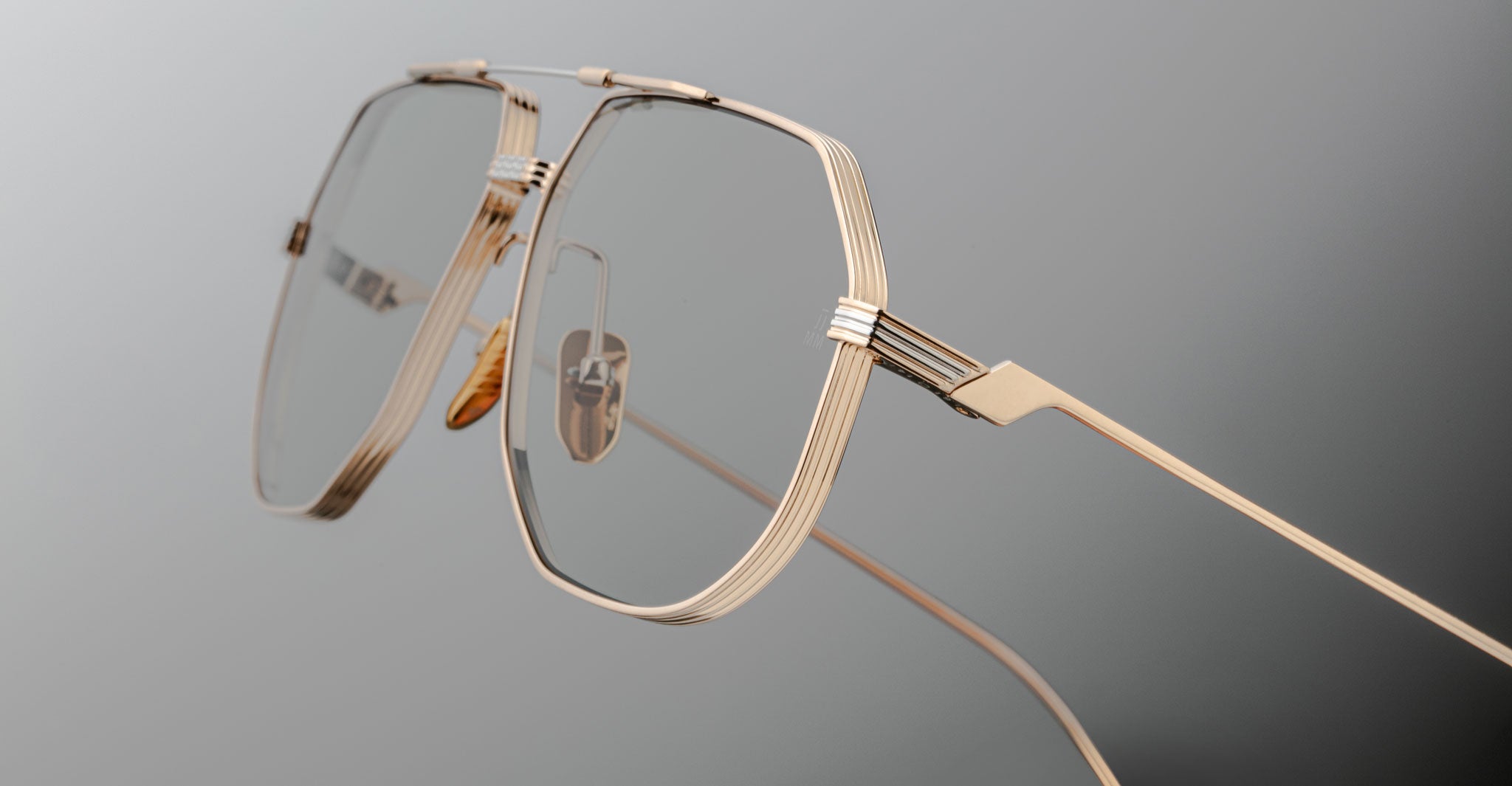 Close-up of Jacques Marie Mage Reynold: stylish eyeglasses with gold rims, a thin titanium frame, and transparent nose pads set against a smooth gradient grey background.