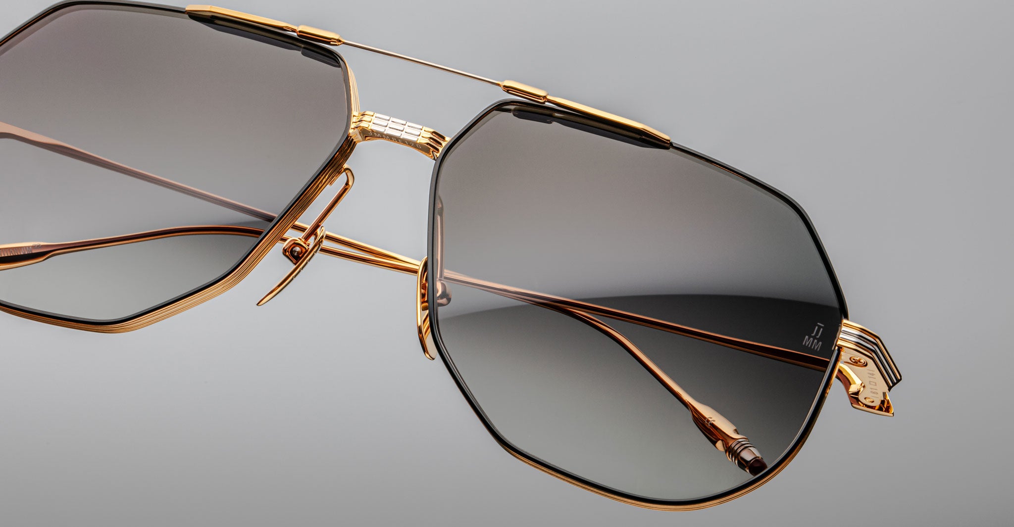 Close-up of Jacques Marie Mage Reynold oversized aviator sunglasses with a gold titanium frame, large dark gradient lenses, and geometric detailing on the arms and nose bridge, set against a light gray background.
