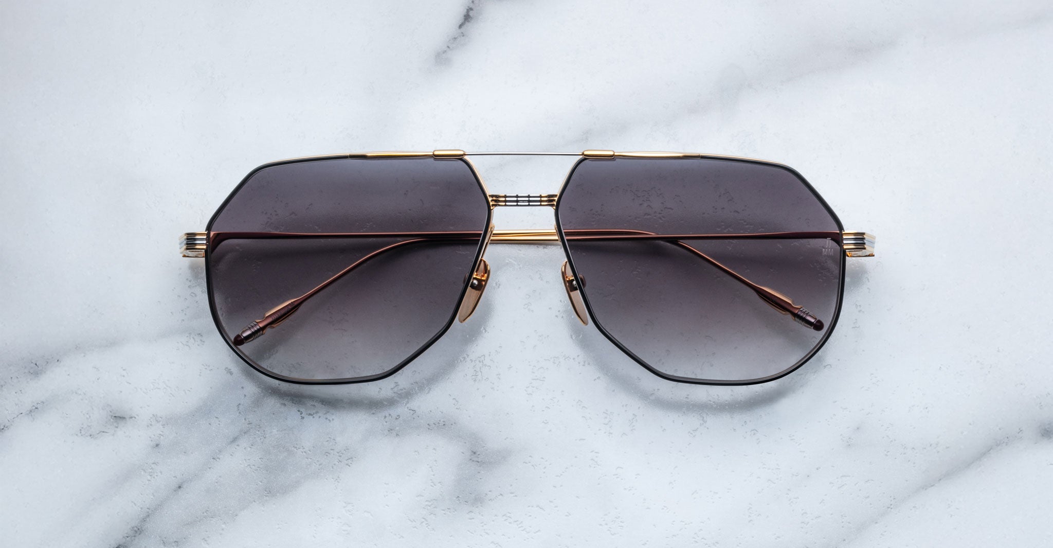The Jacques Marie Mage Reynold features oversized aviator frames in thin gold with dark tinted lenses, styled on a white marble surface—ideal for those who love 70s-inspired eyewear.