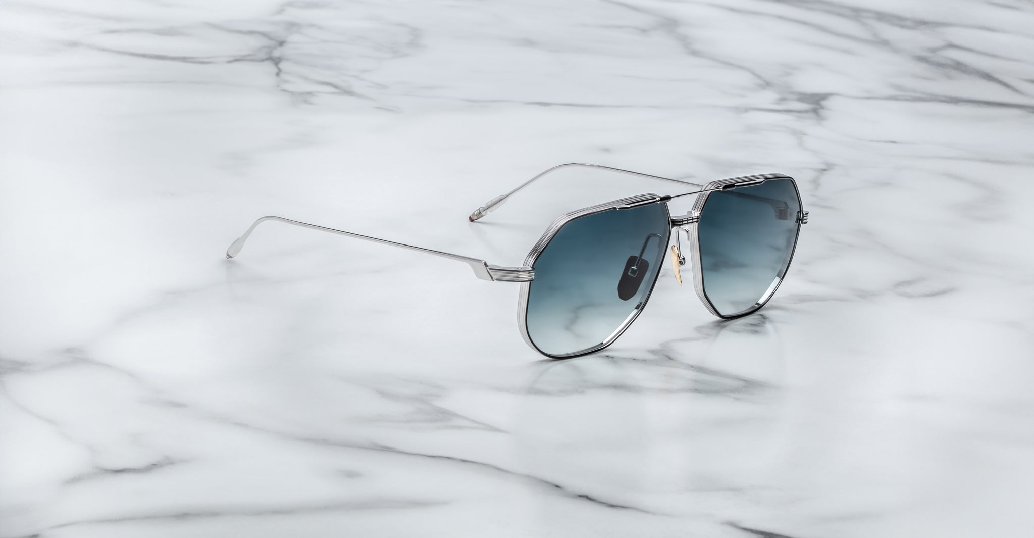 A pair of Jacques Marie Mage Reynold aviator-style sunglasses with silver metal frames and dark tinted lenses rests on a white marble surface with subtle grey veining.