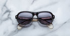 A pair of Jacques Marie Mage Richard Havana, thick-framed sunglasses with blue lenses rests on a white marble surface with grey veins, with faint brand details visible on the inside of the left lens.
