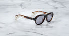 A pair of Jacques Marie Mage Richard Havana, thick-framed sunglasses with blue lenses sits on a white marble surface with grey veins