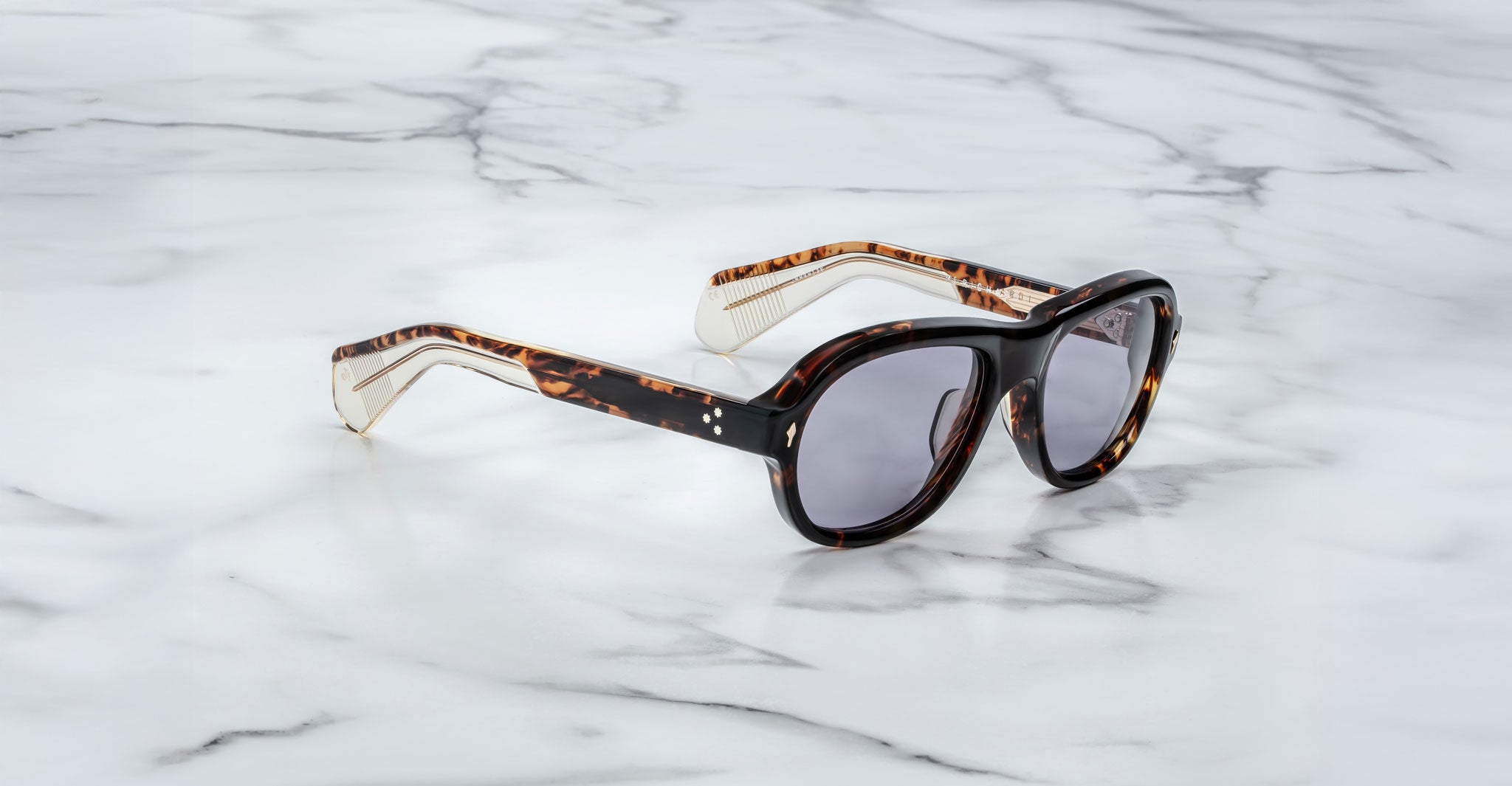 A pair of Jacques Marie Mage Richard Havana, thick-framed sunglasses with blue lenses sits on a white marble surface with grey veins