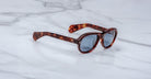 Jacques Marie Mage Richard sunglasses with tortoiseshell thick frames and blue-tinted lenses rest on a white marble surface.