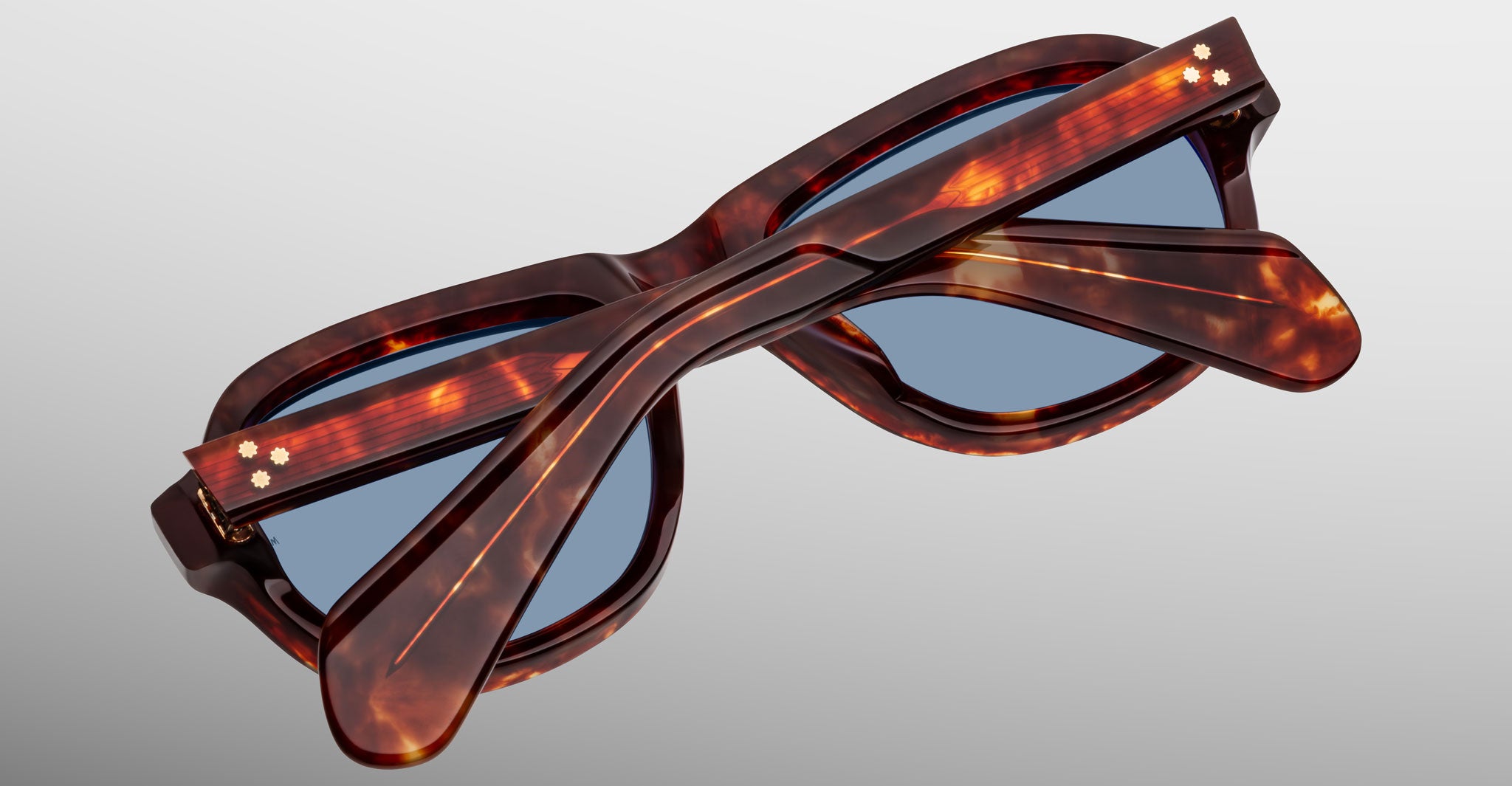The Jacques Marie Mage Richard sunglasses, featuring a tortoiseshell patterned frame and blue lenses, are shown folded closed on a light gray surface with their overlapping arms visible.