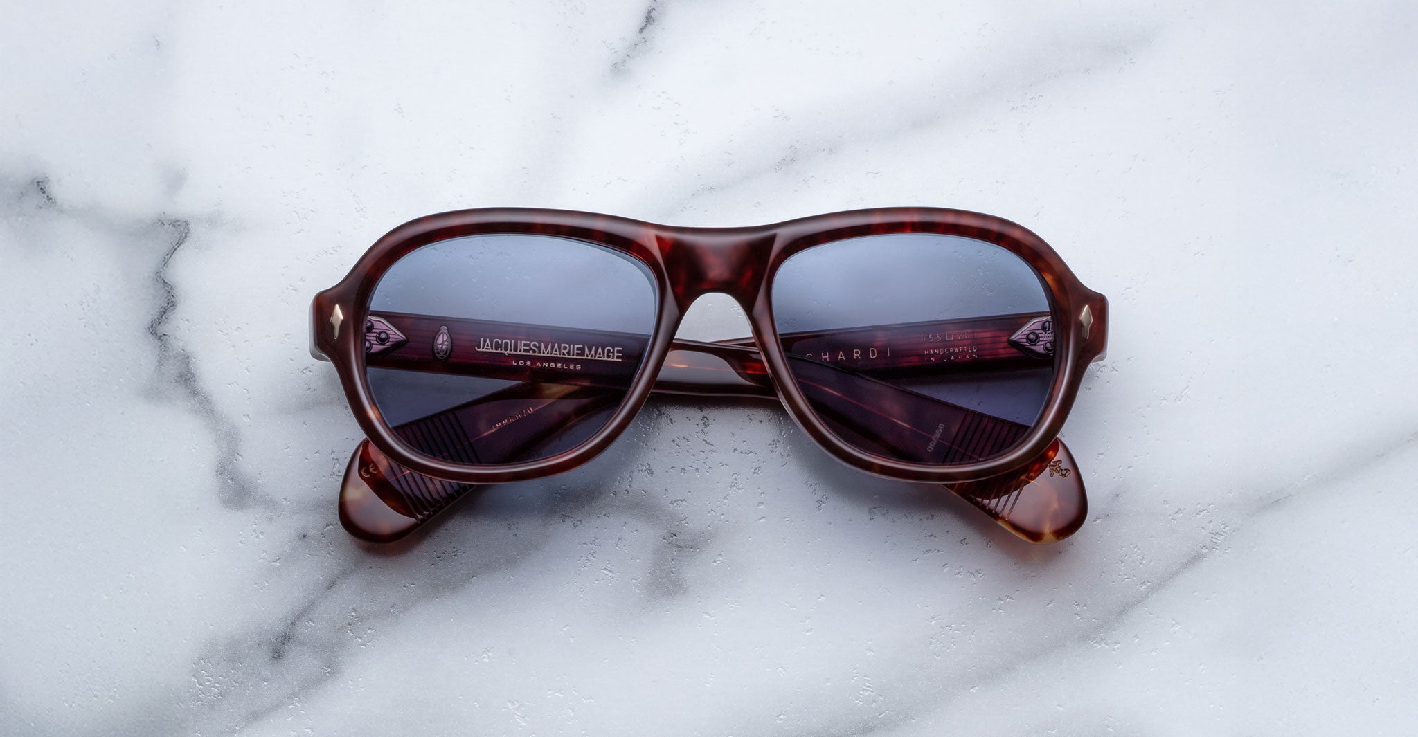 A pair of Jacques Marie Mage Richard sunglasses with dark lenses rests closed on a white marble surface.