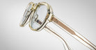 A close-up of Jacques Marie Mage Richard clear round eyeglasses with transparent frames and light gold-tone metal arms, set against a plain light gray background.