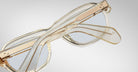 A pair of Jacques Marie Mage Richard eyeglasses in clear, light brown with transparent frames and arms is shown closed on a light gray surface, the arms overlapping each other.