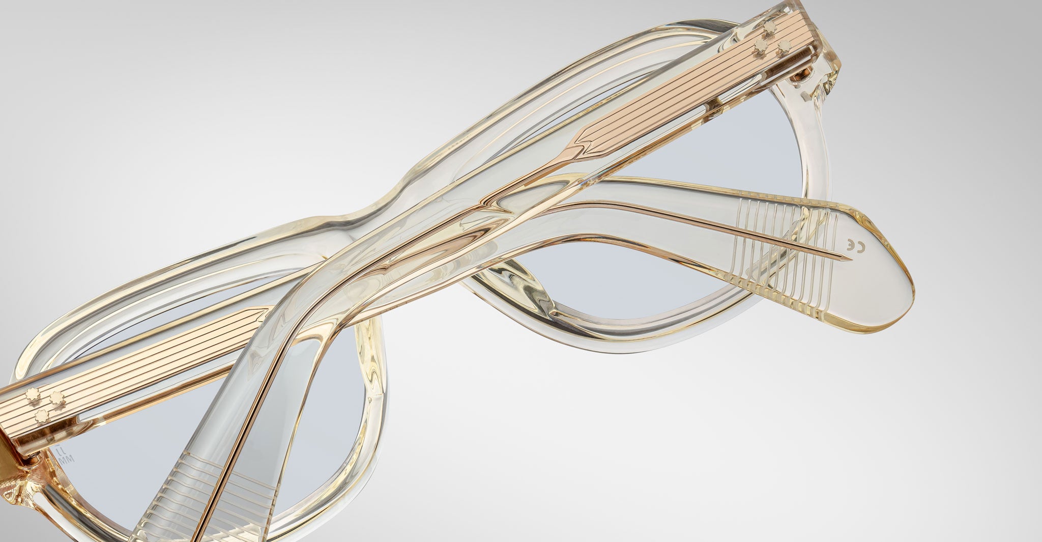 A pair of Jacques Marie Mage Richard eyeglasses in clear, light brown with transparent frames and arms is shown closed on a light gray surface, the arms overlapping each other.