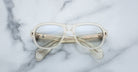 The Jacques Marie Mage Richard eyeglasses, featuring clear frames with a slightly frosted finish, are displayed closed on a white marble surface with the arms crossed behind the lenses.
