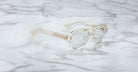 The Jacques Marie Mage Richard eyeglasses, featuring clear thick frames and subtly yellow-tinted temples, rest on a white marble surface with grey veins.
