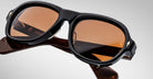 Close-up of Jacques Marie Mage Richard sunglasses with thick, glossy black frames and brown-tinted lenses, resting on their side. The visible arms feature engraved details and text on the inner side.