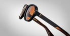 Close-up of Jacques Marie Mage Richard sunglasses featuring black frames, brown-tinted lenses, gold star-shaped accents on the arms, and a visible wood grain pattern on the inner sides, set against a plain gray background.