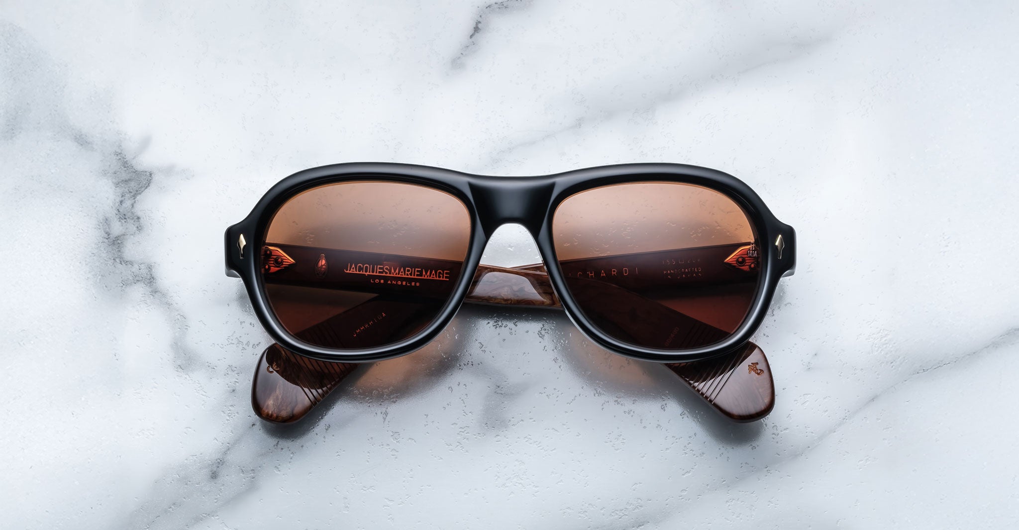 The Jacques Marie Mage Richard sunglasses, featuring black frames and brown-tinted lenses, rest on a white marble surface with partially folded arms and visible reflections on the lenses.