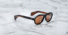 The Jacques Marie Mage Richard sunglasses, featuring black frames, brown-tinted lenses, and wood-textured arm interiors, rest on a white marble surface.