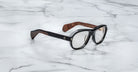 The Jacques Marie Mage Richard eyeglasses, featuring thick black and brown frames, rest on a white marble surface.