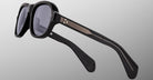 The Jacques Marie Mage Richard sunglasses feature thick black frames, dark lenses, and metallic gold accents with lined temples, displayed on a light gray background.