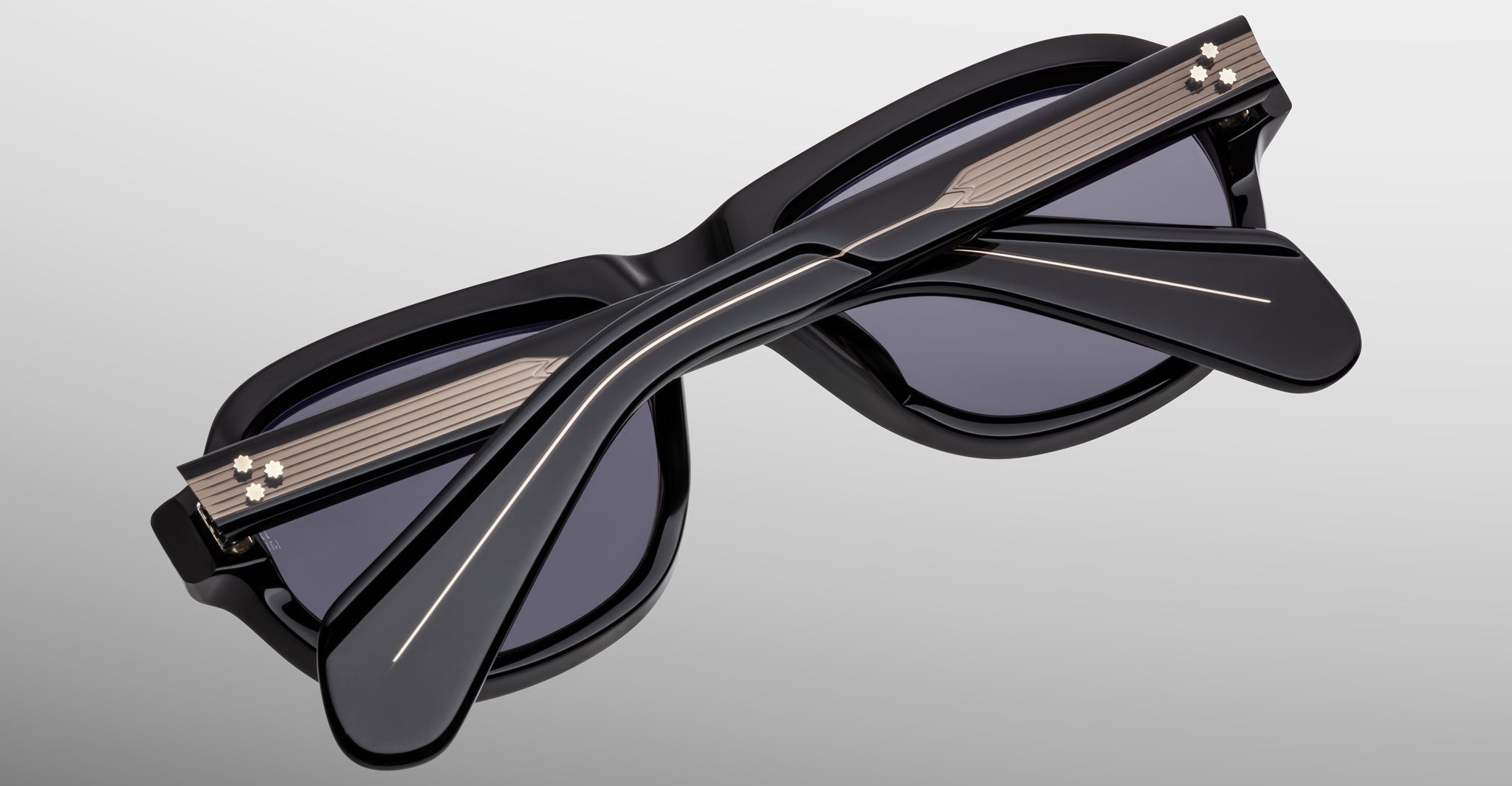 The Jacques Marie Mage Richard sunglasses are stylish black frames with crossed arms over dark lenses, metallic accents near the hinges, set against a light gradient background.