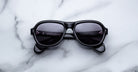 A pair of Jacques Marie Mage Richard black sunglasses with dark lenses sits centrally on a white marble surface, their arms neatly folded.