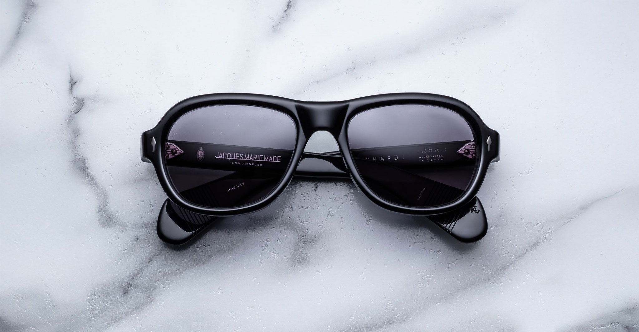 A pair of Jacques Marie Mage Richard black sunglasses with dark lenses sits centrally on a white marble surface, their arms neatly folded.