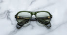 A pair of Jacques Marie Mage Richard green, thick-framed sunglasses with light green lenses rests on a white marble surface with grey veins, with faint brand details visible on the inside of the left lens.