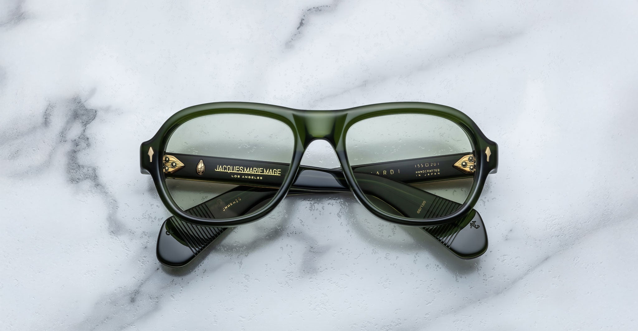 A pair of Jacques Marie Mage Richard green, thick-framed sunglasses with light green lenses rests on a white marble surface with grey veins, with faint brand details visible on the inside of the left lens.