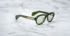 A pair of Jacques Marie Mage Richard green, thick-framed sunglasses with light green lenses sits on a white marble surface with grey veins