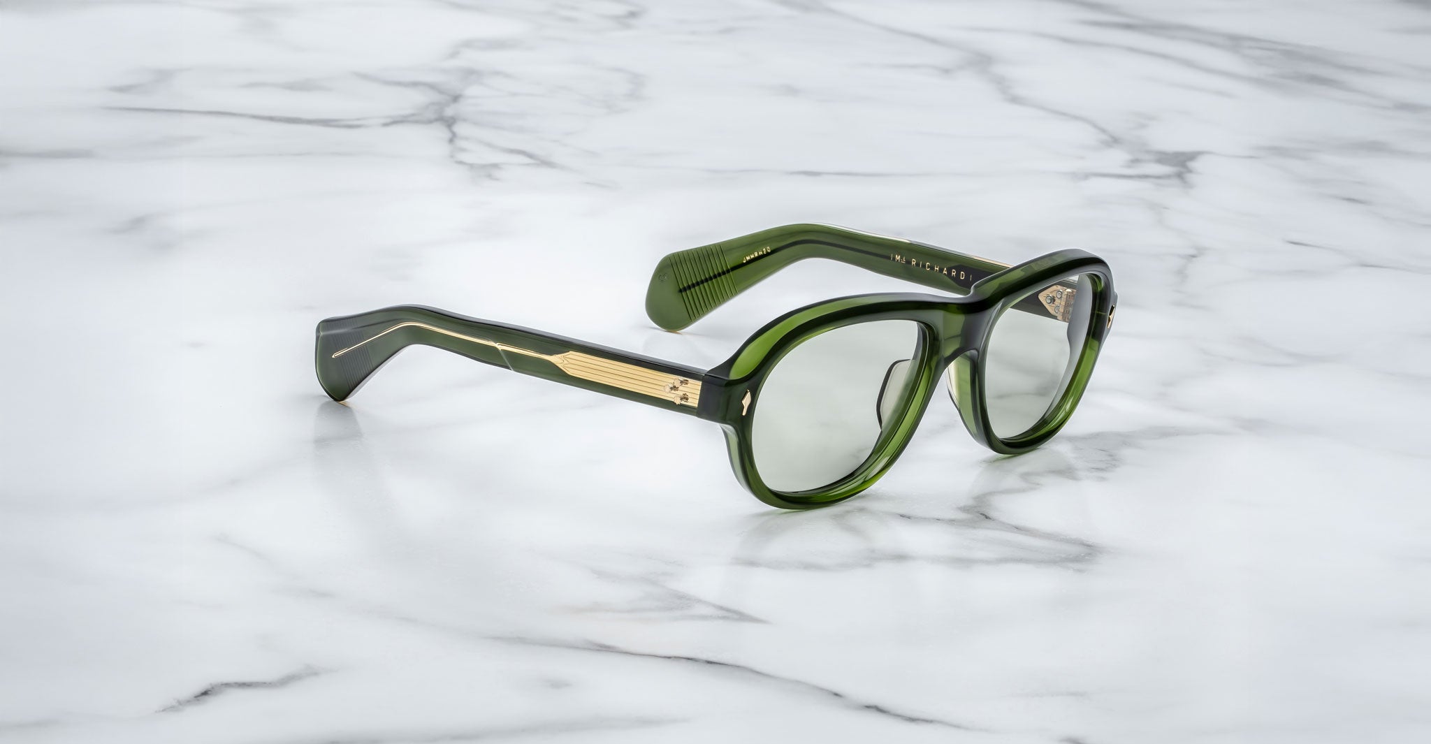 A pair of Jacques Marie Mage Richard green, thick-framed sunglasses with light green lenses sits on a white marble surface with grey veins