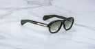 A pair of Jacques Marie Mage Richard black sunglasses with dark lenses and gold detailing on the arms rests on a white marble surface.