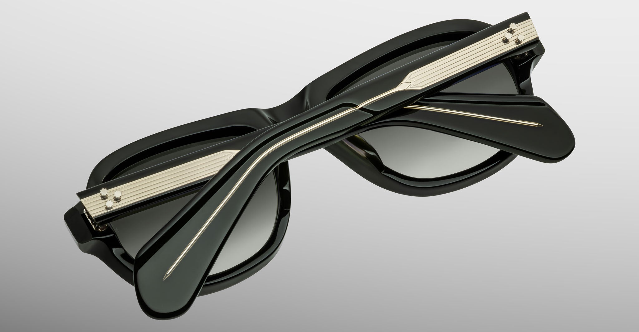 A close-up of the Jacques Marie Mage Richard sunglasses folded, highlighting gold accents on the arms and a sleek black modern design against a light gray background.