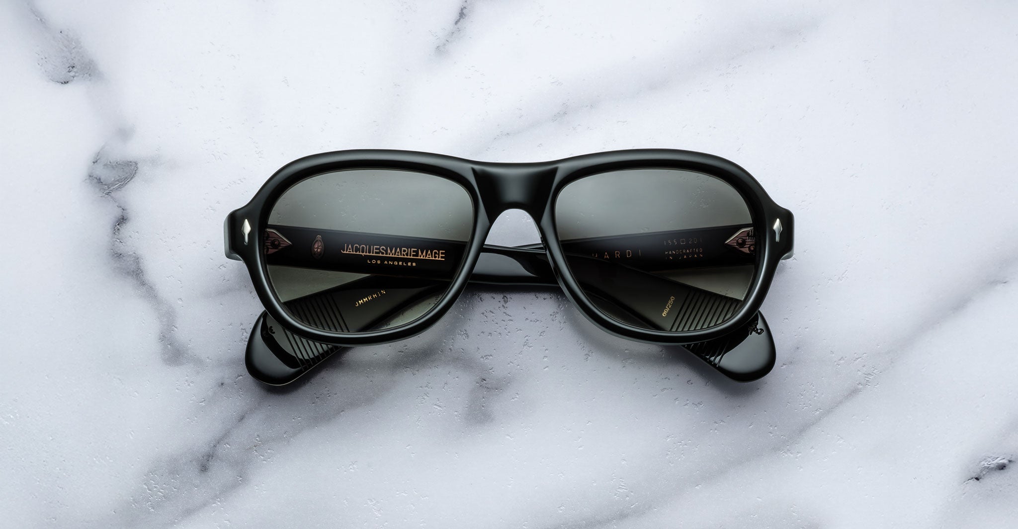 A pair of Jacques Marie Mage Richard black sunglasses with dark lenses and thick frames lies upside down on a white marble surface, displaying gold text on the inner arms.