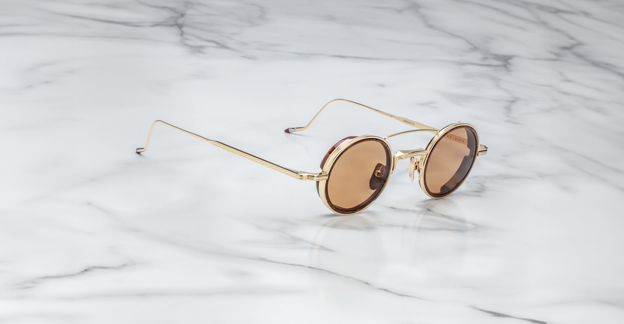 The Jacques Marie Mage Ringo sunglasses feature gold frames and brown-tinted lenses, resting at an angle on a white marble surface with grey veining, highlighting their thin arms and modern design.