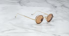 The Jacques Marie Mage Ringo sunglasses feature gold frames and brown-tinted lenses, resting at an angle on a white marble surface with grey veining, highlighting their thin arms and modern design.